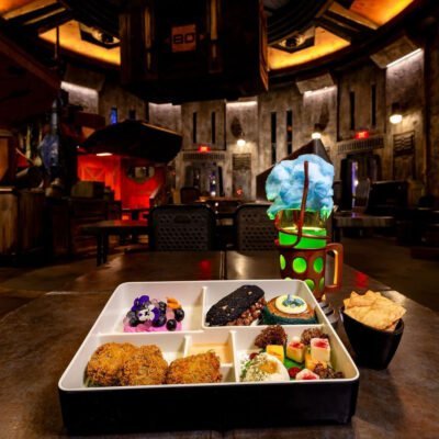 A bento-style food tray with various dishes including fried items, a dessert with blueberries and edible flowers, and chips, is placed on a table. Beside the tray is a drink topped with blue cotton candy in a futuristic mug. The background shows a dimly lit, sci-fi themed restaurant interior.