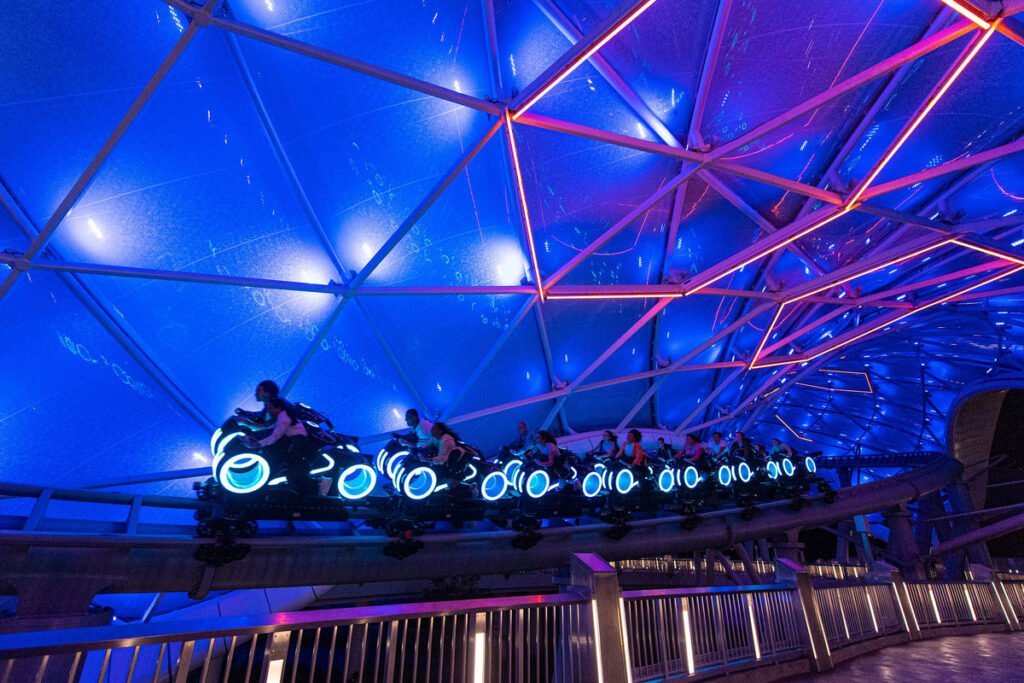 People riding a futuristic roller coaster with glowing blue lights under a geometrically structured ceiling with blue and red lighting.