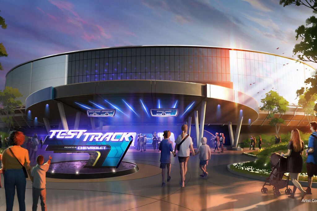 Entrance to the Test Track attraction at a theme park, with people walking towards the illuminated sign and modern building structure in the background.