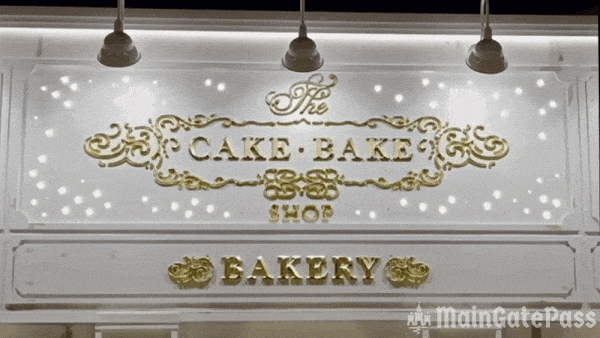Signage of a bakery, featuring the text "The CAKE BAKE SHOP" in decorative gold lettering, with lights illuminating the background.