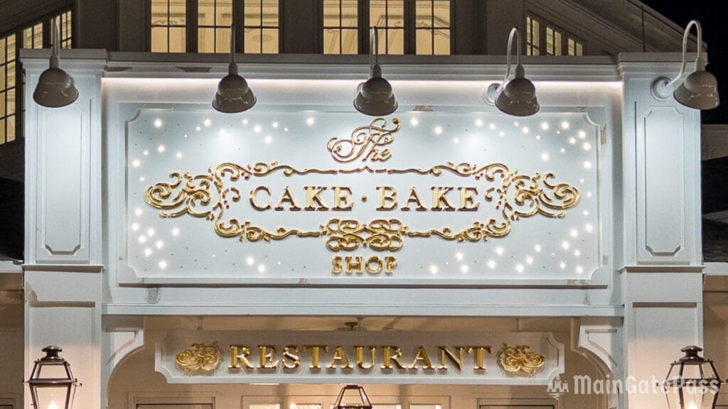 Signage for "The Cake Bake Shop" restaurant, featuring ornate gold lettering and decorative elements, illuminated against a white background.