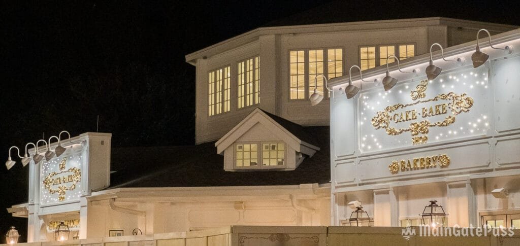 A well-lit bakery sign reading "The Cake Bake Shop Bakery" is displayed on a white building at night, featuring ornate gold lettering and decorative elements. Multiple light fixtures are attached above the sign, illuminating the surroundings.
