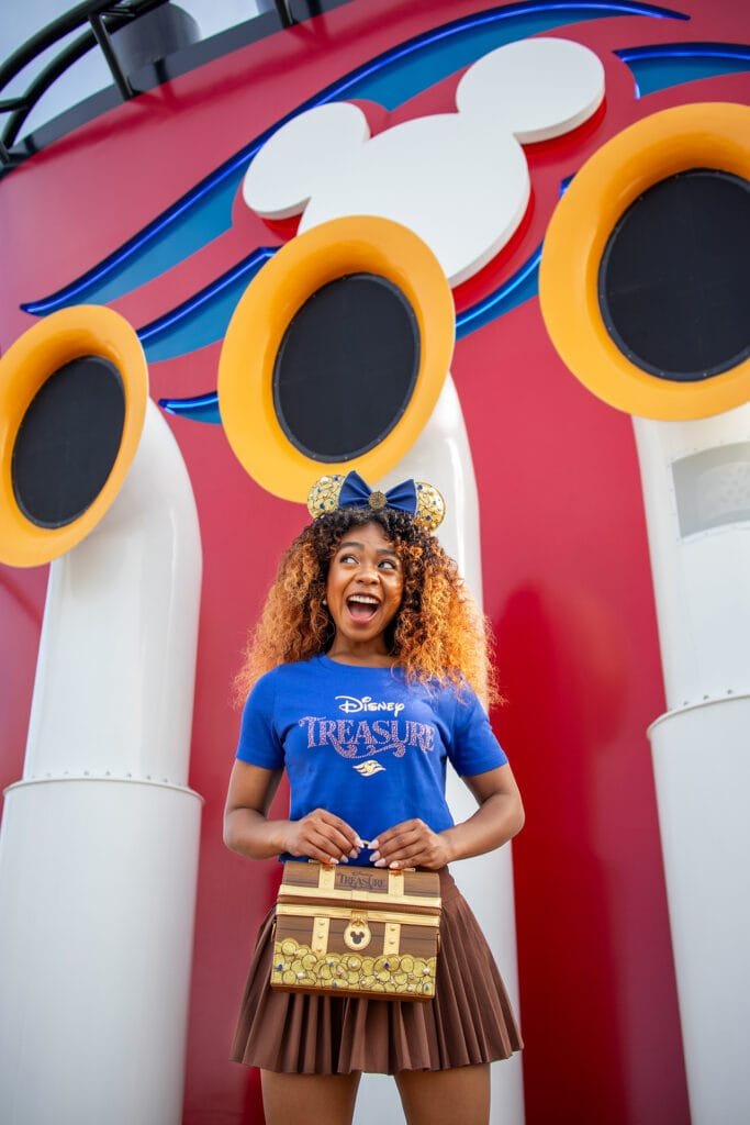 A smiling person with curly hair wears a blue Disney-themed shirt and gold mouse ear headphones, holding a decorative treasure chest bag, in front of a colorful backdrop featuring large yellow and black speakers.