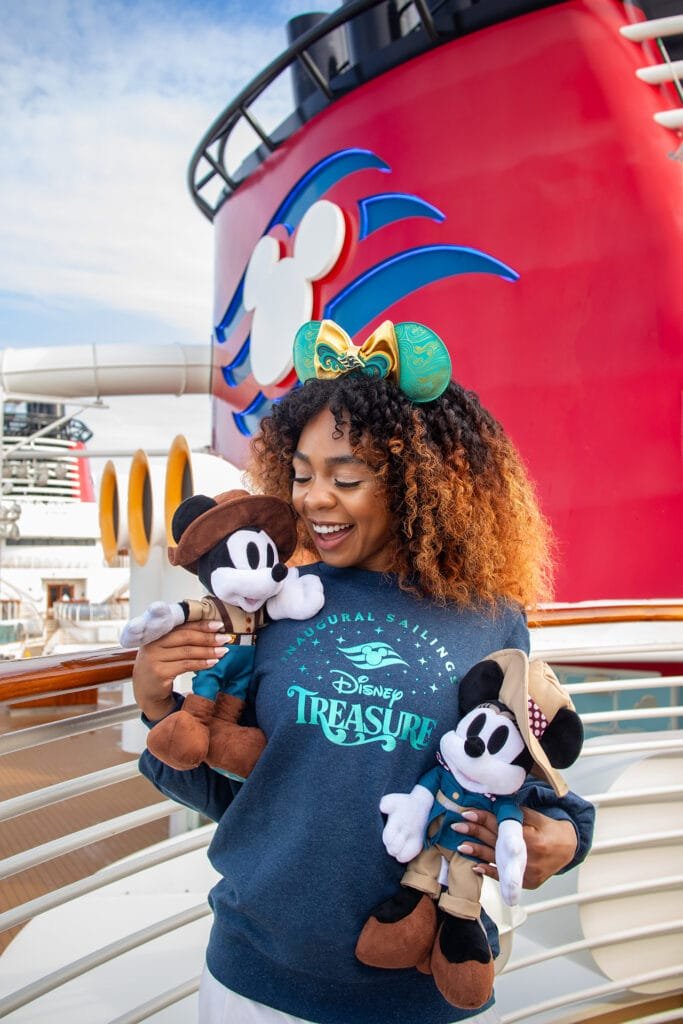A person with curly hair wearing a dark sweatshirt holds two Mickey Mouse plush toys, one dressed as an explorer and the other in a brown hat. The background features a cruise ship with a red hull and a stylized mouse ear design.