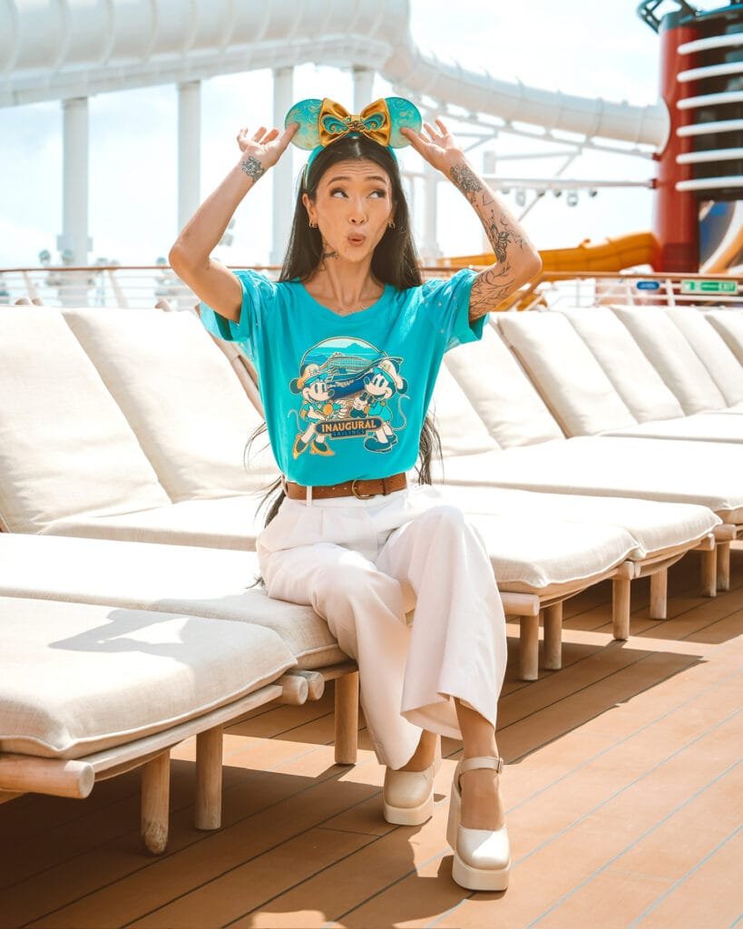 A woman with long hair and tattoos sits on a lounge chair, wearing a bright turquoise t-shirt featuring cartoon characters and white wide-legged pants. She has a large bow headband on her head and is playfully making a funny face while raising her hands. The background shows a sunny outdoor area with lounge chairs and a slide.