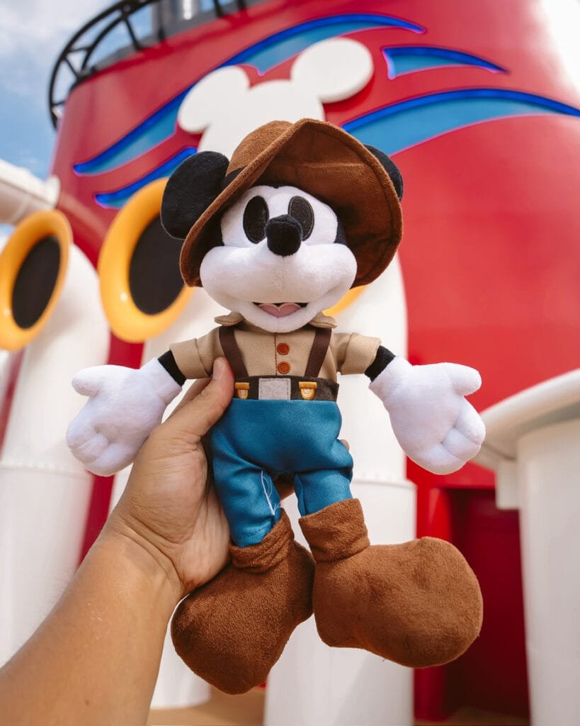 A hand holds a plush Mickey Mouse toy dressed in a brown hat, beige shirt, and blue pants, with a colorful cruise ship backdrop featuring large yellow portholes and a red and blue design.