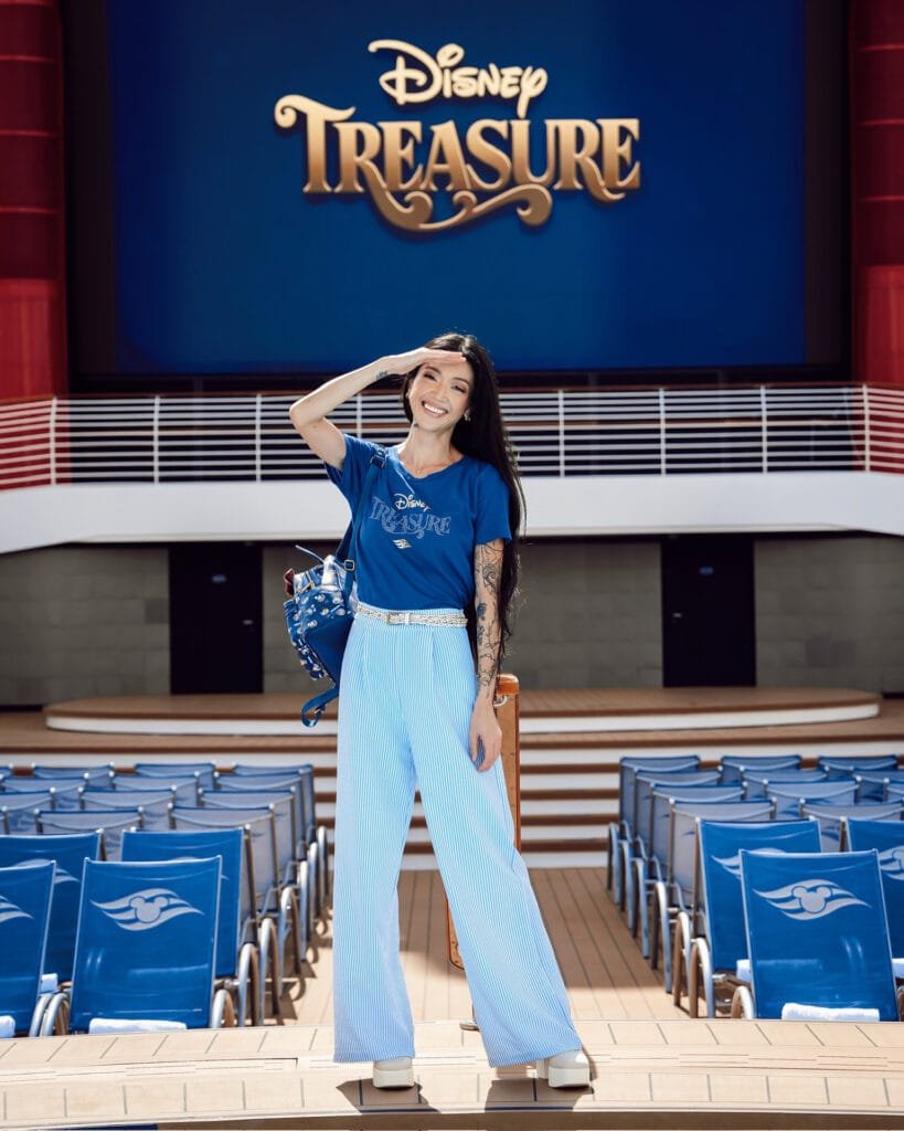 A woman with long dark hair stands on a cruise ship deck, smiling and waving. She is wearing a blue Disney Treasure t-shirt and wide-legged light blue striped pants. Behind her, there is a large sign displaying "Disney Treasure" against a blue backdrop, with empty lounge chairs visible.