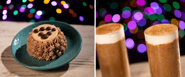 A chocolate cake with decorative icing and chocolate candies on a teal plate beside two tall glasses of brown foam-topped beverages, set against a background of colorful, blurred lights.