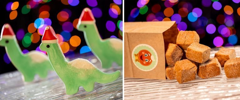 Three green dinosaur-shaped cookies wearing Santa hats are in the foreground, with a colorful, blurred background. In the background, there's a small, open carton with a label featuring a goldfish, containing brown, sugar-like cubes.