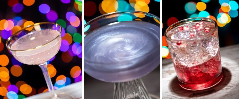 Three colorful cocktails are displayed against a blurred background of vibrant lights. The left cocktail is a sparkling pink drink in a coupe glass, the middle is a shimmering purple drink in a wide-rimmed glass, and the right is a red drink with ice and garnishes in a short glass.