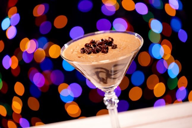 A chocolate dessert in a martini glass, topped with chocolate shavings, against a blurred background of colorful holiday lights.