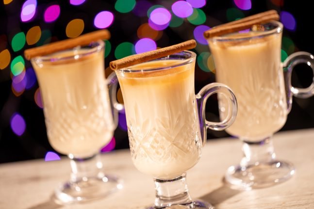 Three glass mugs filled with a creamy beverage, each topped with a cinnamon stick, set against a colorful, blurred background of lights.