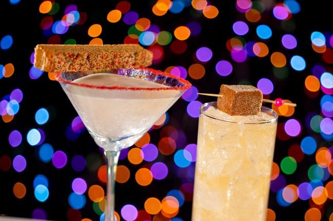A close-up of two cocktails against a colorful bokeh background with lights. The left drink is a martini decorated with a festive cookie and rimmed with red sugar, while the right drink is a tall glass filled with ice and a garnish of a brown sugar cube.