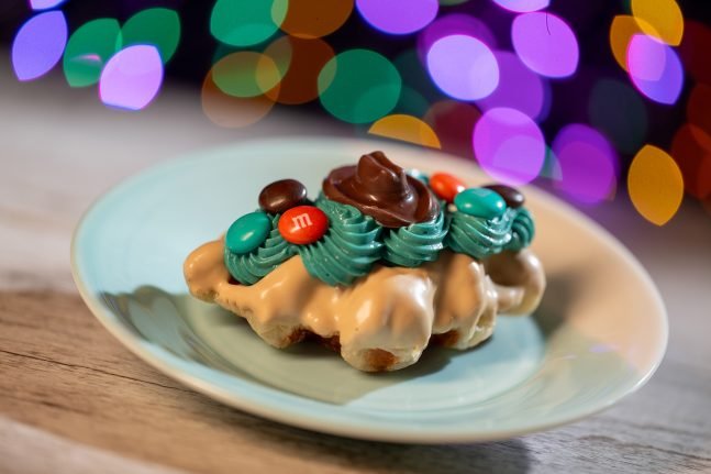 A decorated pastry topped with teal frosting, chocolate, and colorful candies, placed on a light blue plate, with a blurred background of multicolored lights.