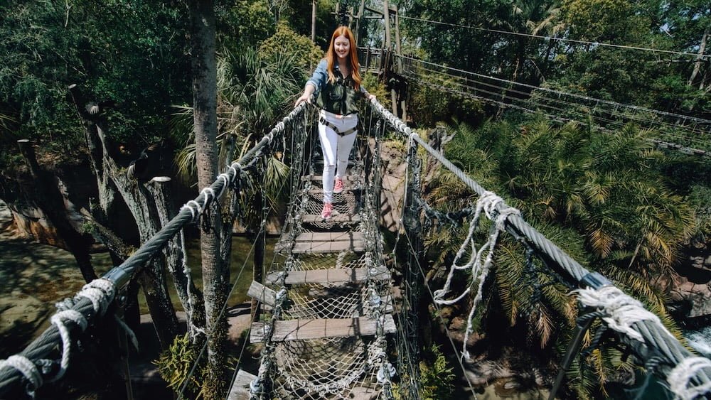A woman with long red hair walks confidently on a suspension bridge surrounded by lush greenery and palm trees.