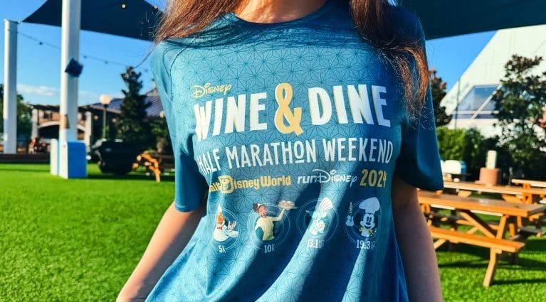 Person wearing a blue Disney t-shirt that reads "WINE & DINE HALF MARATHON WEEKEND 2021," featuring images of Disney characters and race distances.