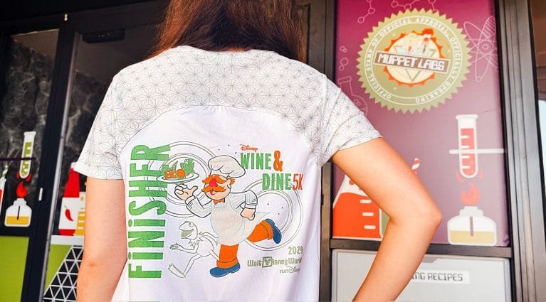 A person is shown from the back wearing a white shirt with a graphic design featuring a chef, the text "FINISHER," and details of the "Disney Wine & Dine 5K" event from 2022. The background includes a colorful sign with a "Muppet Labs" theme.