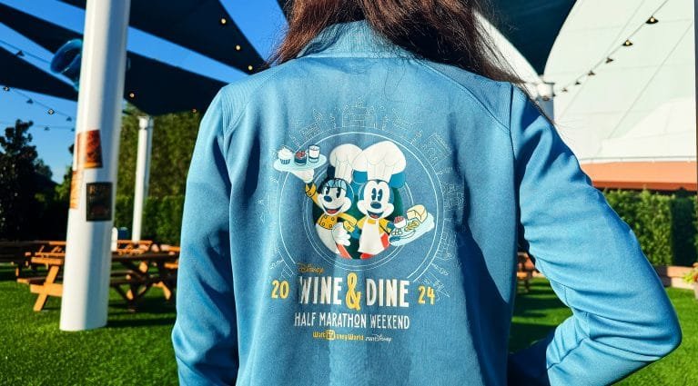 A person is wearing a blue jacket featuring a graphic on the back with two animated characters resembling chefs, holding desserts. The design includes the text "WINE & DINE" and "HALF MARATHON WEEKEND 2024" along with the Disney logo. The setting is outdoors with greenery and outdoor seating visible.