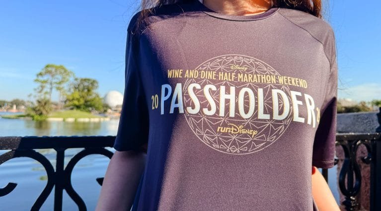 A close-up of a person wearing a black shirt with the text "20 PASSHOLDER" and "WINE AND DINE HALF MARATHON WEEKEND" printed in yellow, with a scenic view of a lake and trees in the background.