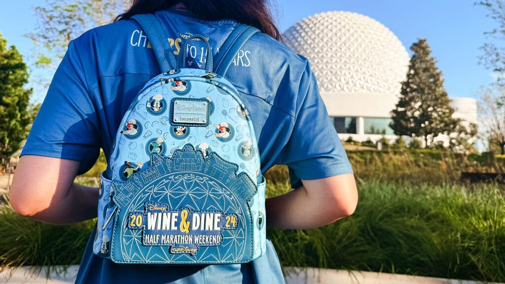 A person wearing a blue shirt is seen from behind, showcasing a light blue backpack decorated with Disney characters and text that reads "Disney WINE & DINE HALF MARATHON WEEKEND 2024." In the background, there is a large white geodesic dome structure and some greenery.