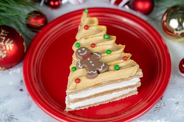A slice of cake shaped like a Christmas tree, decorated with frosting in green and red, topped with a gingerbread man cookie, on a red plate, surrounded by holiday ornaments.