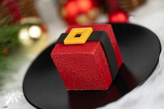 A red gift-shaped dessert with a black ribbon and yellow buckle sits on a black plate, surrounded by festive decor.
