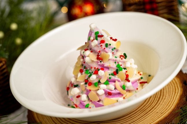 A decorative dessert shaped like a Christmas tree, featuring pink and white frosting, topped with marshmallows, almond slices, and colorful sprinkles, served in a white bowl on a wooden surface with festive decorations in the background.