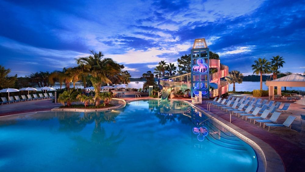 A brightly lit outdoor swimming pool surrounded by palm trees and lounge chairs, with a water slide in the background and a vibrant evening sky.