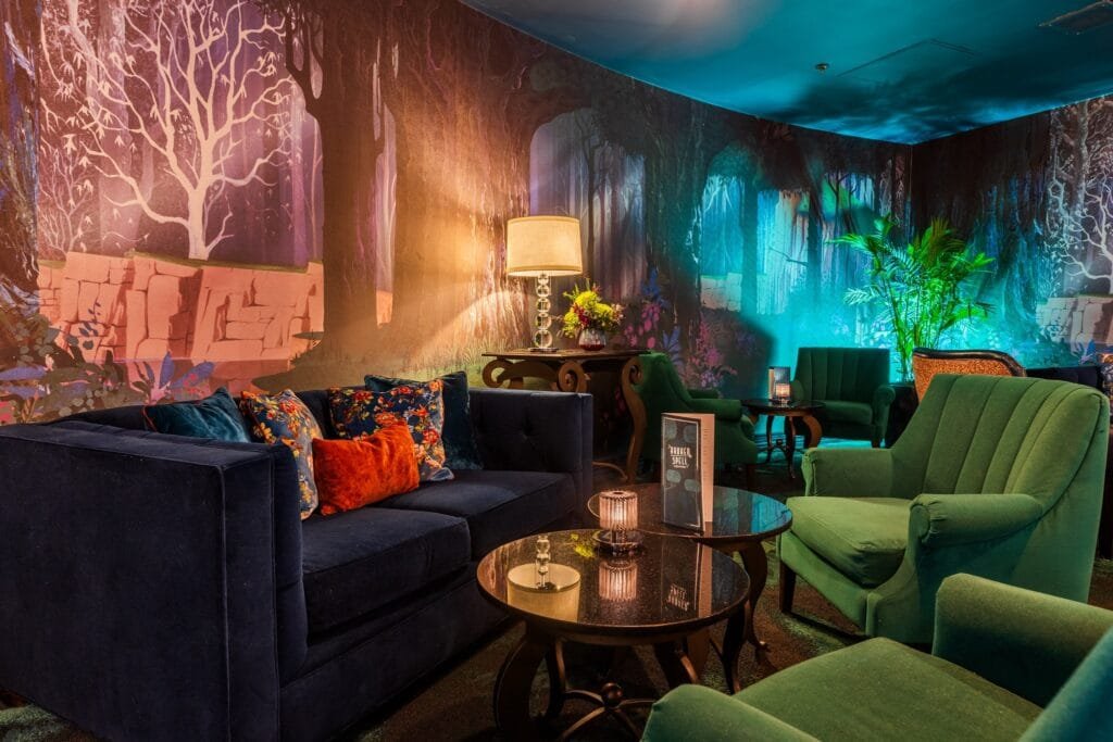 Cozy lounge area featuring a dark blue velvet sofa with colorful patterned cushions, green velvet chairs, and round tables. The background has a vibrant forest-themed mural with trees and plants, illuminated by soft lighting from a nearby lamp.