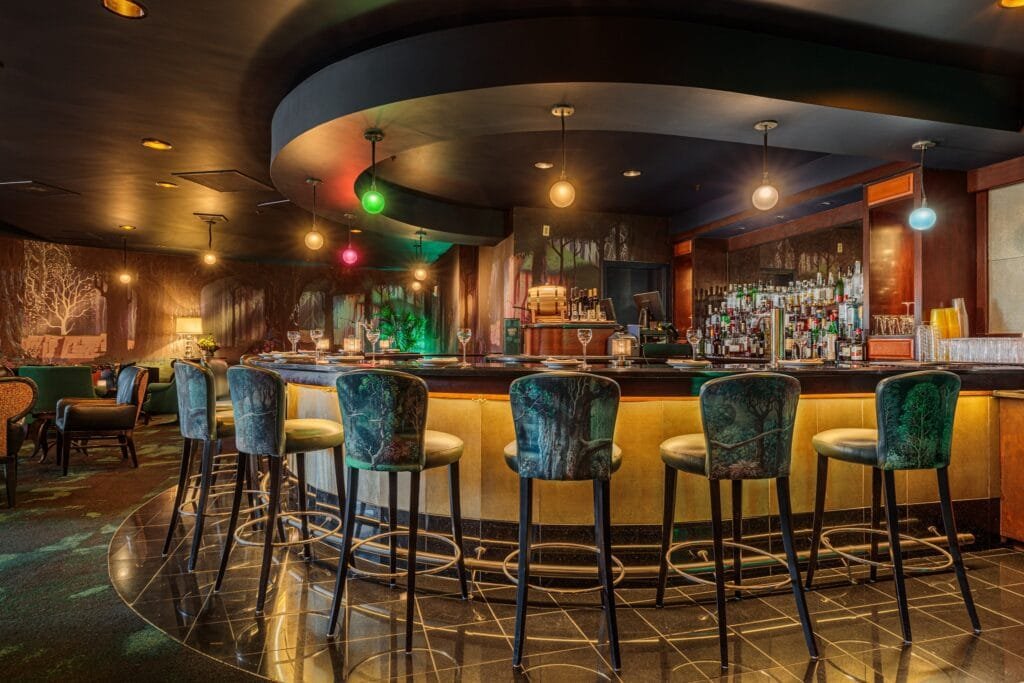 A stylish bar interior featuring a curved counter with high stools, illuminated by pendant lights in various colors. The bar is adorned with a collection of liquor bottles behind it, and the walls are decorated with forest-themed murals. The floor has a shiny black tile finish, creating an elegant atmosphere.