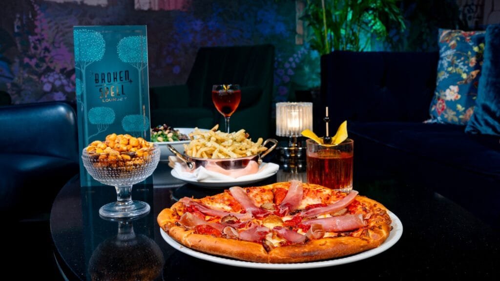 A table setting featuring a pizza topped with meats, a bowl of snacks, a plate of French fries, a salad, a cocktail, and a glass of drink, with the "BROKEN SPELL LOUNGE" sign in the background.