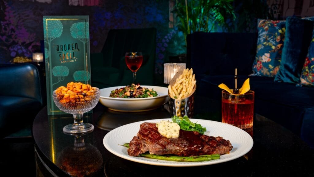 A plate of steak with melted butter on top rests on green beans, accompanied by a salad and fries in metal containers, a bowl of nuts, and cocktails on a dark table. A menu card for "Broken Spell Lounge" is visible in the background.
