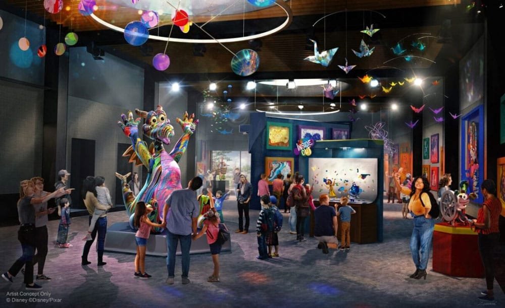 A colorful exhibition space filled with visitors, showcasing vibrant artwork and a central sculpture of a whimsical creature. The room features various interactive displays and hanging decorations, with children and adults engaged in the displays around them.