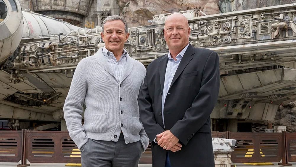 Two men are standing in front of a large, detailed spaceship replica. One man is wearing a light gray sweater over a collared shirt, and the other is in a black suit with a light blue shirt. They are smiling and posing for the photo.