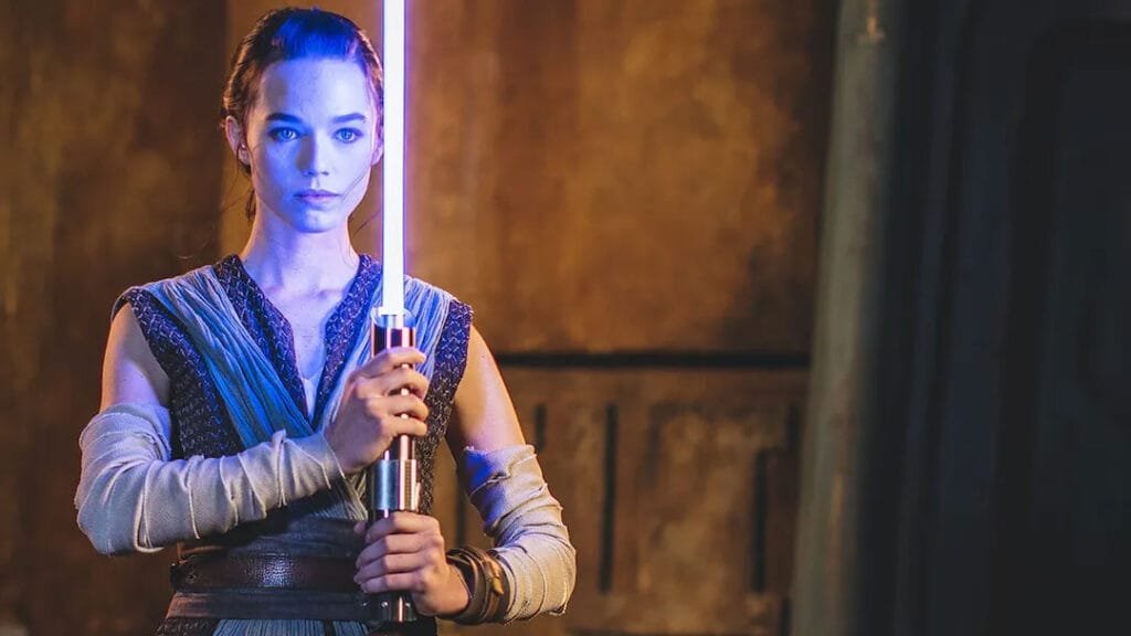 A woman in a fantasy costume holds a glowing lightsaber, with a serious expression, set against a textured, warm-toned background.