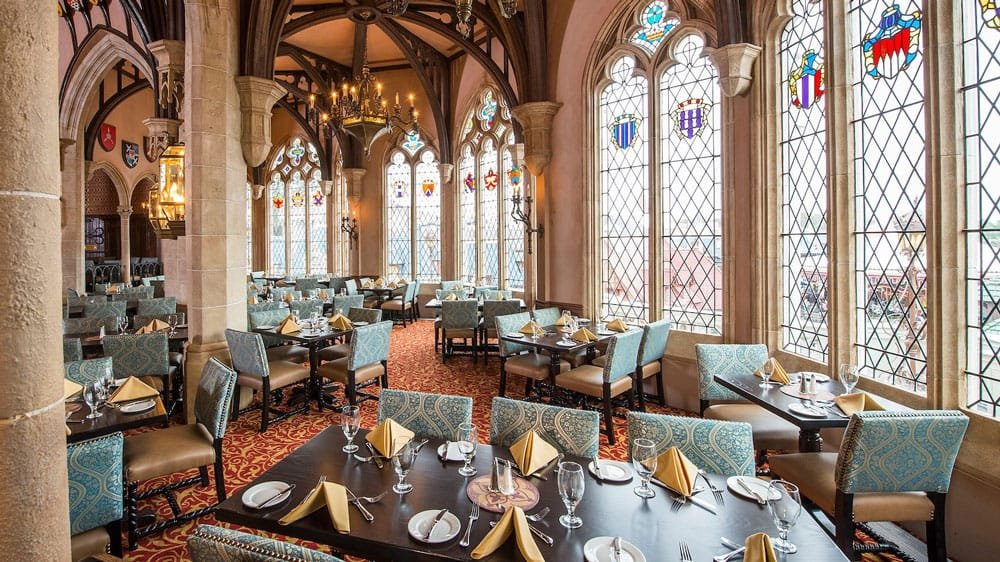 A grand dining room featuring elegant tables set with fine tableware, surrounded by high ceilings and large stained glass windows. The decor includes ornate chandeliers and patterned upholstery on chairs, creating a luxurious atmosphere.