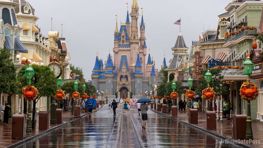 A rainy scene of a quiet main street in a theme park, lined with colorful decorations and trees, leading to a large, ornate castle in the background. People walk under umbrellas along the wet pavement.