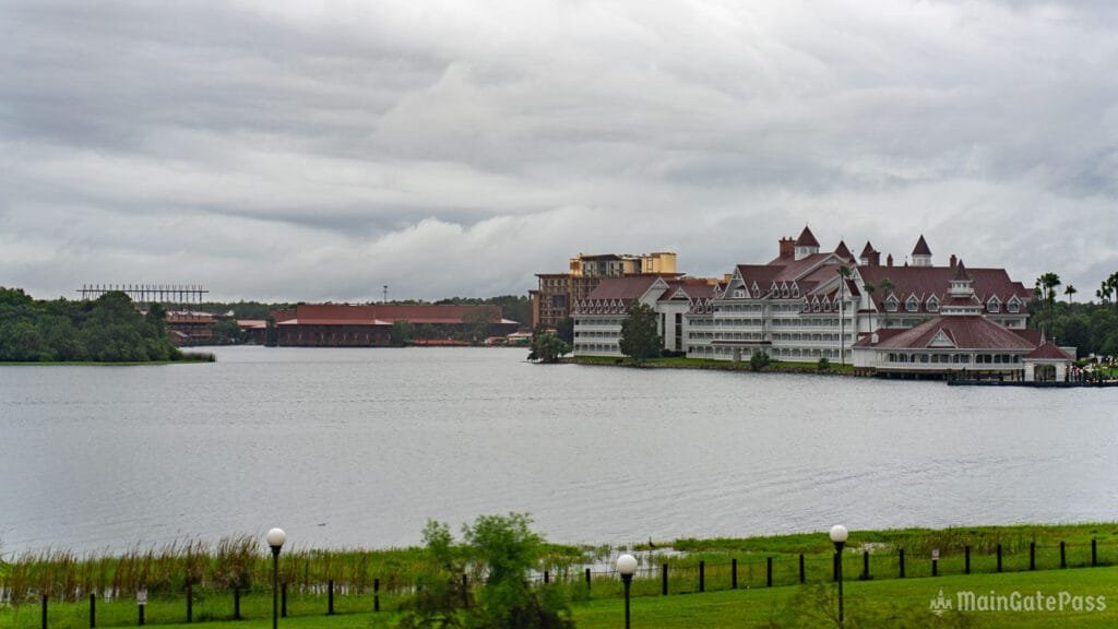A large lake reflecting a cloudy sky, with a white and red-roofed hotel on the right and several trees and grass along the shore. In the background, another building and a partially completed structure are visible.