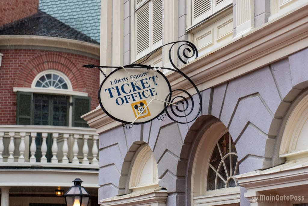 Sign for the Liberty Square Ticket Office, hanging from a decorative bracket on a building, with a background of a brick structure and green shutters.