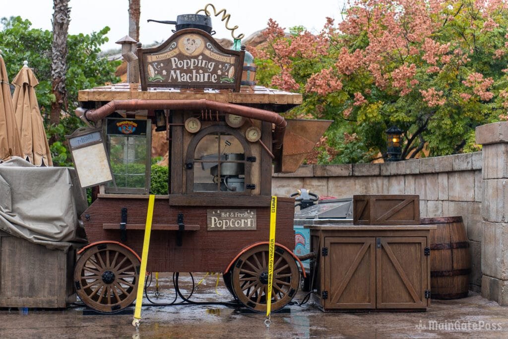 A vintage popcorn cart named "Popping Machine" with a rustic design, featuring wooden wheels and a glass display for popcorn. It is adorned with signs advertising hot and fresh popcorn, and is surrounded by trees and decorative elements in a rainy outdoor setting.