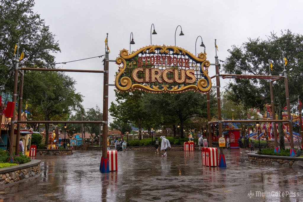 Entrance to Storybook Circus, featuring a large, colorful sign and a rainy, reflective ground with people in raincoats.