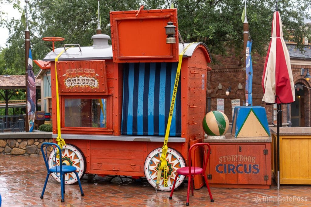 A colorful food stand resembling a circus wagon, featuring a sign that reads "Storybook Circus Shacks," with blue and red chairs in front, a beach ball, and various decorative elements including striped curtains and flags. The scene is set on a brick pavement with greenery in the background.