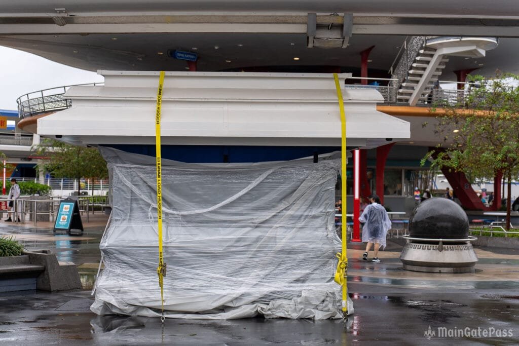 A covered booth secured with yellow straps is shown in a wet outdoor area, with a couple of people in rain gear walking in the background and a large black spherical structure nearby.