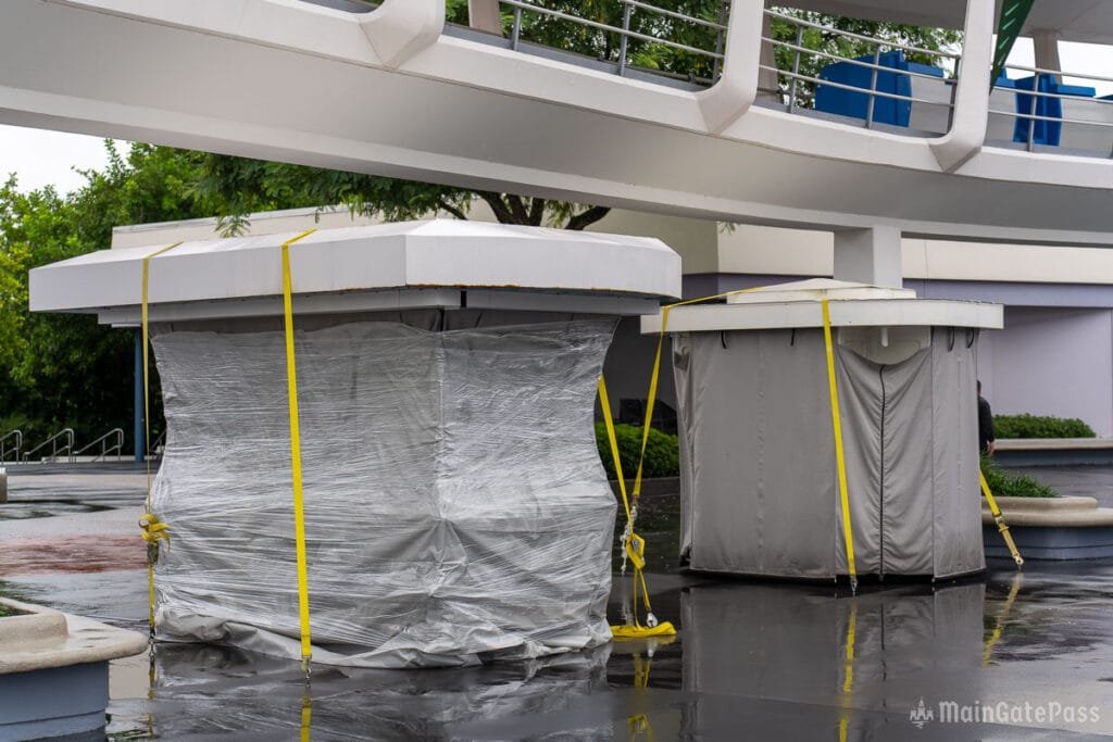 Two square structures covered in plastic wrap and gray fabric, secured with yellow straps, are positioned under a walkway. The ground is wet, reflecting the surroundings, and there are trees and a building in the background.