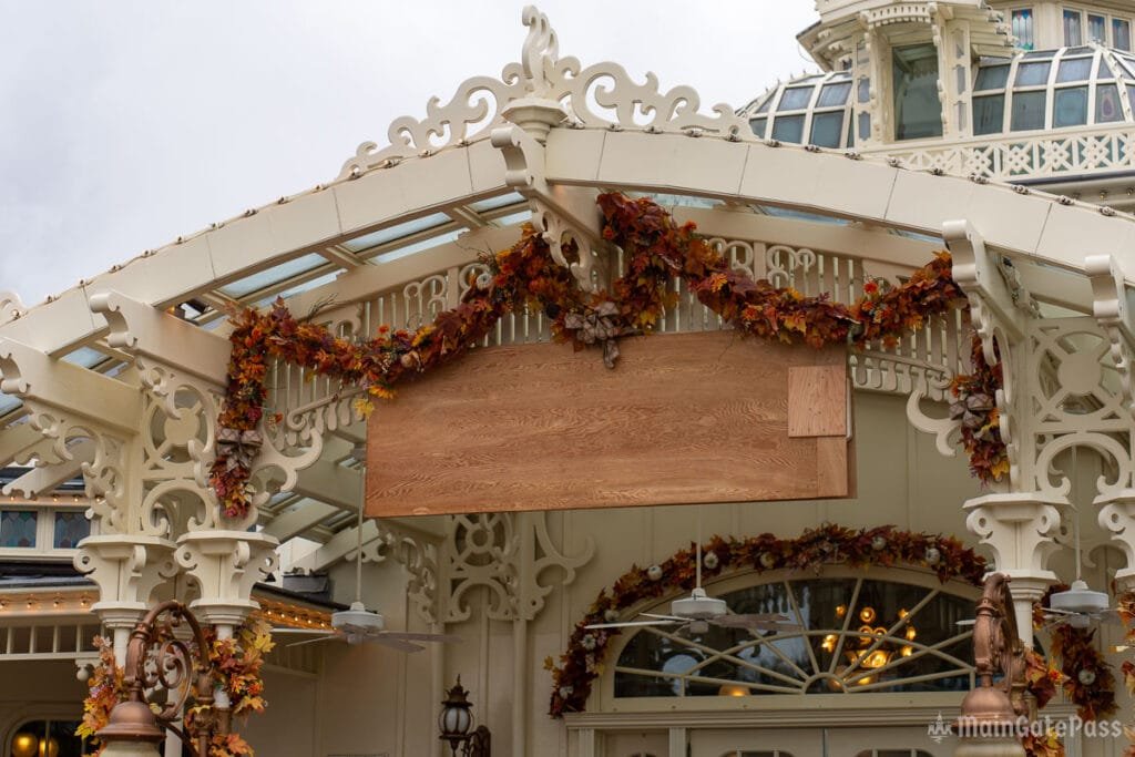 A decorative entrance featuring intricate white lattice work and a wooden sign covered, adorned with fall leaves and flowers. Above the entrance, glass panels allow light through, and there are hanging lights and ceiling fans visible.