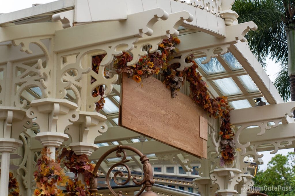 A decorative pavilion structure with intricate white filigree, a blank wooden sign hanging in the center, and autumn leaves draped along the edges.