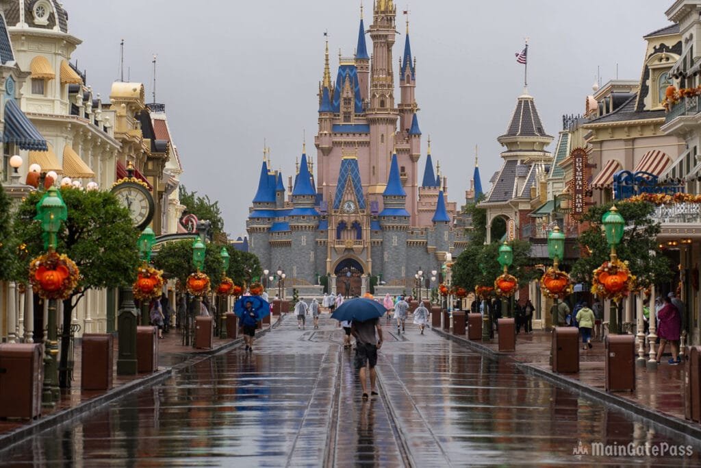 A rainy day at a theme park with a view of a fairytale castle in the background. People walk down a wet pathway lined with trees and decorative lanterns. Some individuals are carrying umbrellas and wearing raincoats.