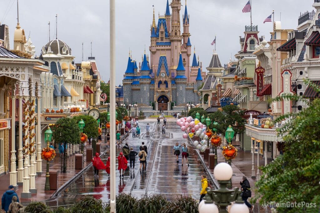 A view of a rainy Main Street at a theme park, featuring colorful storefronts and decorations, with a castle in the background and visitors walking down the wet path. Balloons are on display, and some people wear raincoats.