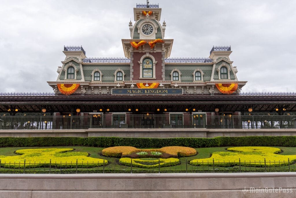 Facade of the Magic Kingdom entrance featuring decorative landscaping in the shape of Mickey Mouse, under a cloudy sky.
