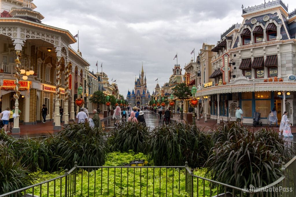 Empty street scene at a theme park filled with shops, featuring lush green plants in the foreground and a castle in the background under a cloudy sky.